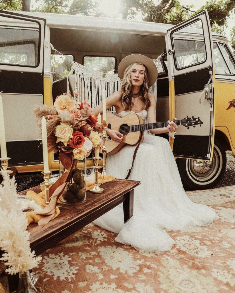 bride in her tulle wedding dress sitting  by the camping van while playing a guitar