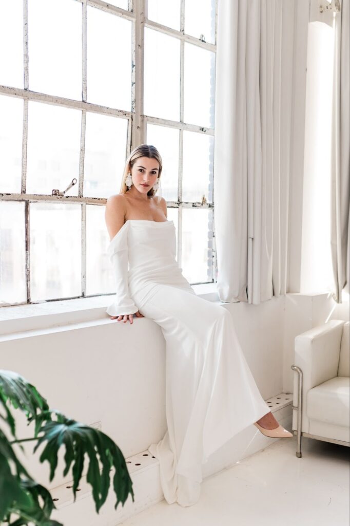 a woman wearing an off-shoulder wedding dress sitting by the window